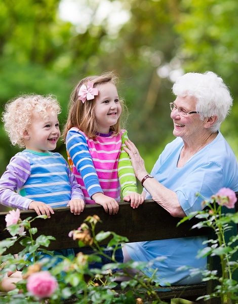 Happy senior lady playing with little boy and girl in blooming rose garden. Grandmother with grand children sitting on a bench in summer park with beautiful flowers. Kids gardening with grandparent.