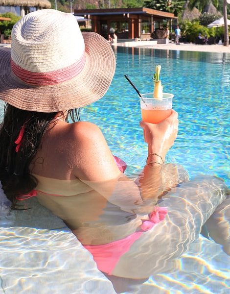 Beautiful woman using hat and bikini enjoy a tropical resort sitting front pool and drinking cocktail back view.