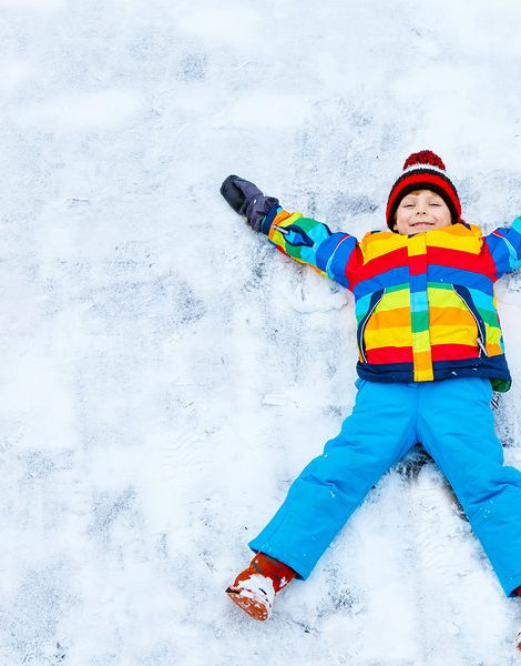 Cute little kid boy in colorful winter clothes making snow angel, laying down on snow. Active outdoors leisure with children in winter. Happy child