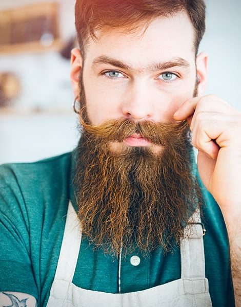 Closeup portrait of handsome young man with beard in white apron touching his moustache