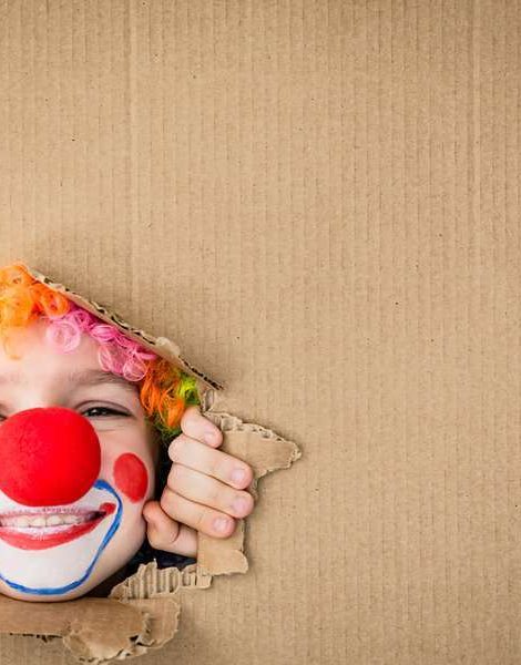 Funny kid clown looking through hole on cardboard. Child playing at home. 1 April Fool's day concept. Copy space.