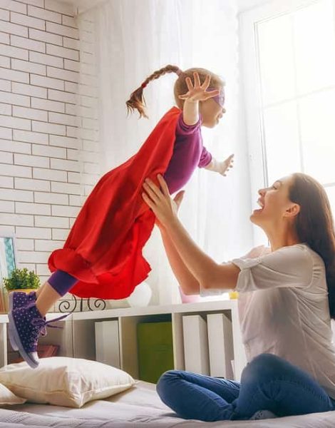 Mother and her child girl playing together. Girl in an Superman's costume. The child having fun and jumping on the bed.