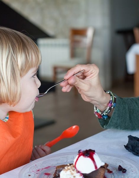 green sweater woman feeding little kid orange bib with metal spoon a piece of chocolate cake with vanilla ice cream at restaurant